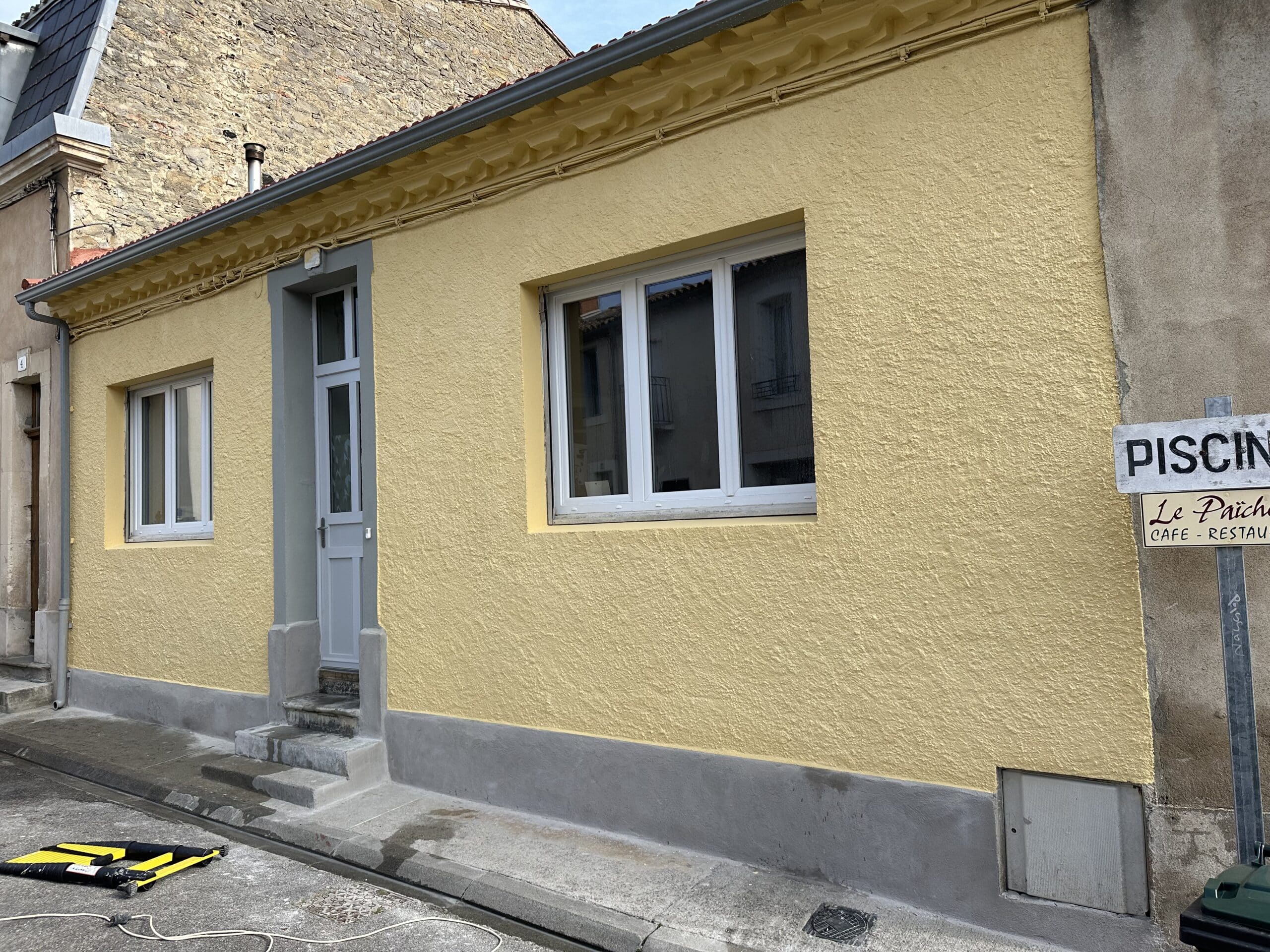 Façade crépie jaune vif d'une maison de ville. Corniche détaillée, porte grise et fenêtres PVC. Signalisation "PISCINE Le Païche CAFE - RESTAU".