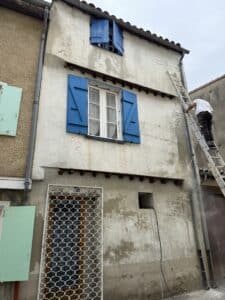 Ouvrier sur une échelle nettoyant à haute pression une façade décrépite aux volets bleus. Stuc blanc, gouttière, grille ornementale.