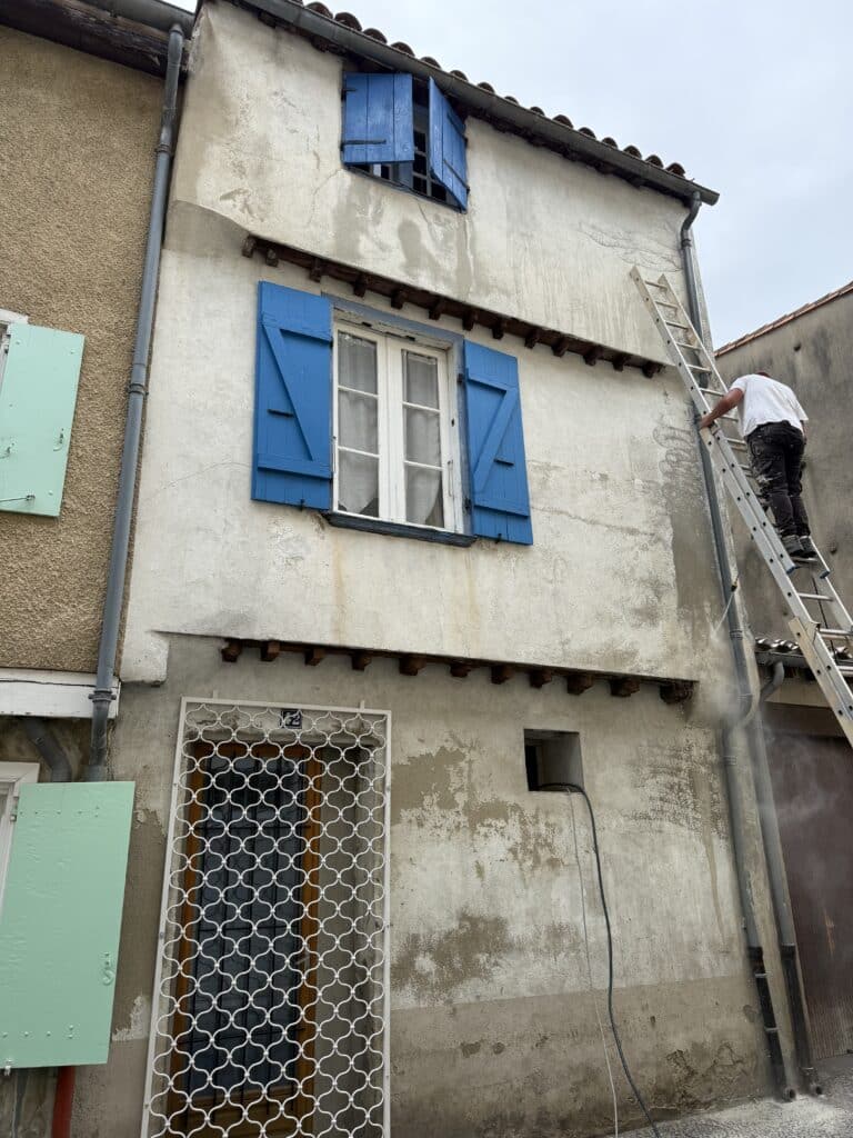 Façade ancienne : Nettoyage haute pression et volets bleus Ouvrier sur une échelle nettoyant à haute pression une façade décrépite aux volets bleus. Stuc blanc, gouttière, grille ornementale.