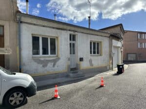 Façade de maison en stuc blanc avec fenêtres PVC. Les traces de ravalement jaunes et grises dominent. Deux cônes et panneau "PISCINE" sur la rue.
