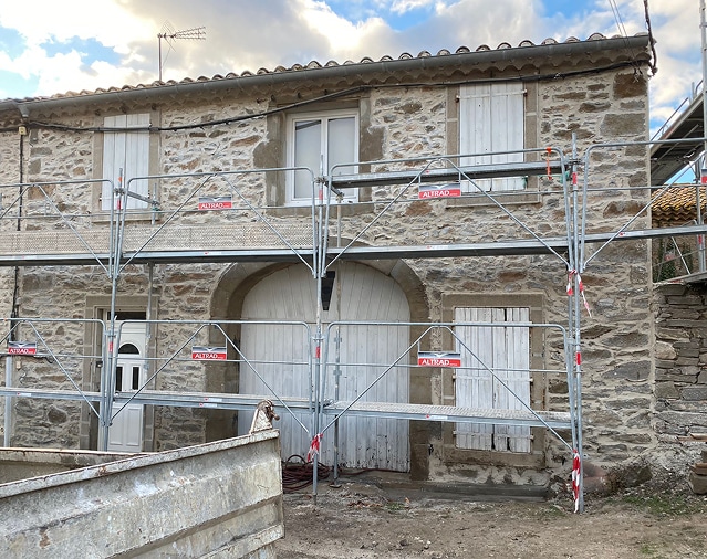 Façade en pierre de maison ancienne en rénovation, couverte par un échafaudage ALTRAD. Porte cintrée et volets bois blancs.
