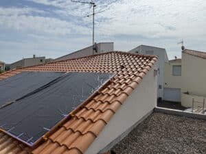 Toit en tuiles vernies rouges sous un ciel nuageux. Une bâche noire de protection est fixée pour des travaux solaires. Terrasse en gravier au premier plan.