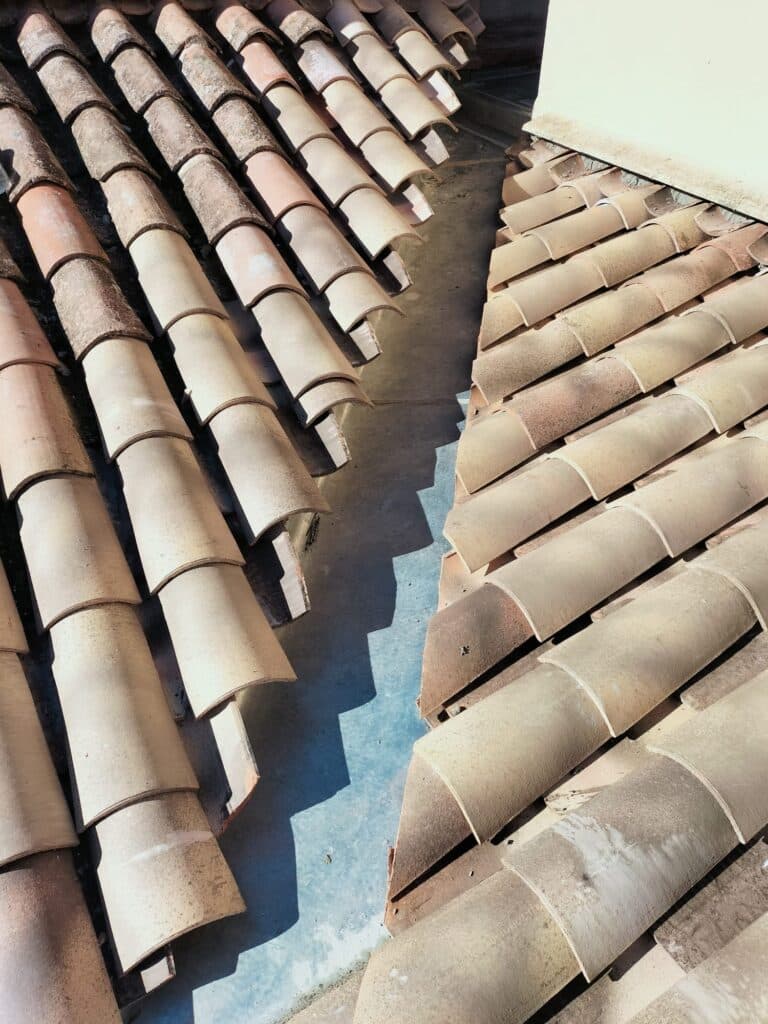 Tuiles de toit canal en terre cuite : détail de la construction Detail en gros plan de tuiles canal en terre cuite superposees, montrant la jonction d'une noue ombragee avec un mur blanc. Lumiere vive.