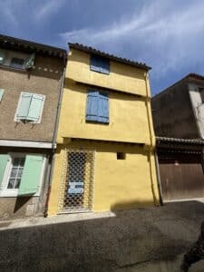 Façade jaune vibrante avec volets bleus dans une ruelle provençale. Grille blanche et panneau "AUDE rénovation" pour le rez-de-chaussée. Ciel bleu.