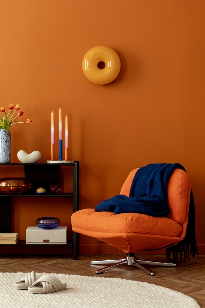 Fauteuil orange design et déco mur terracotta moderne. Fauteuil orange moderne et plaid bleu contre un mur terracotta. Déco design avec étagère noire, bougies multicolores et art mural donut jaune.