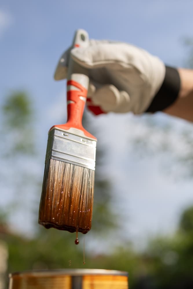 Pinceau lasure bois extérieur : application simple et protection. Pinceau chargé de lasure marron, une goutte s'échappant vers le pot. Main gantée et ciel bleu : application extérieure de protection bois.