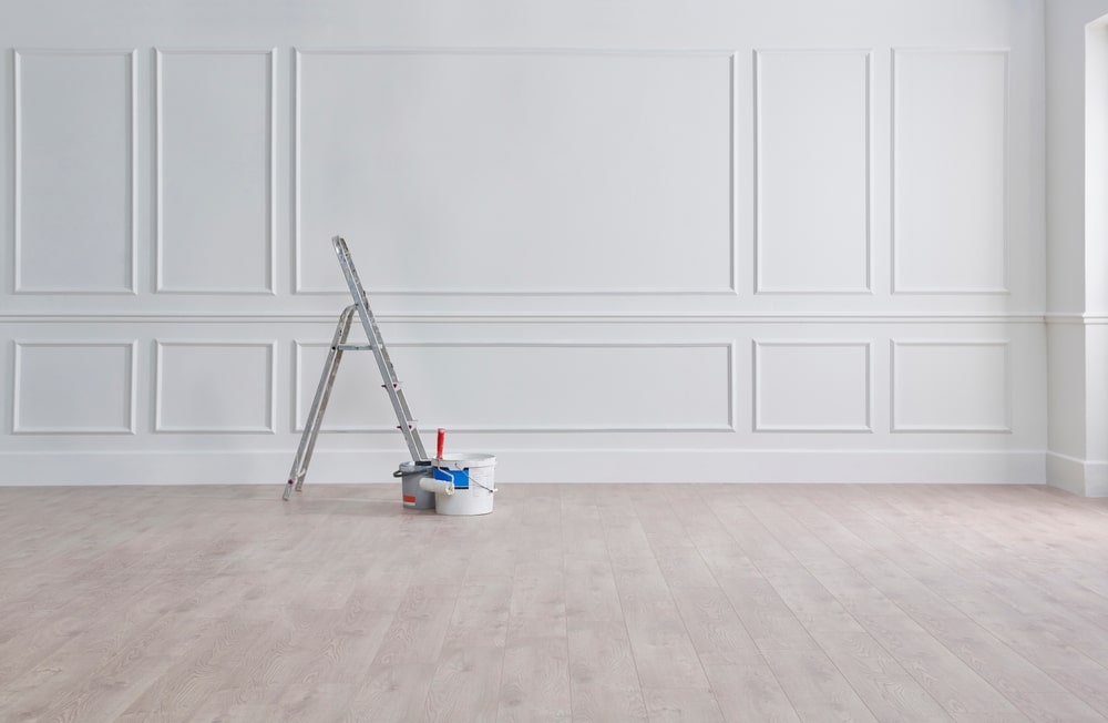 Mur blanc classique à moulures et boiseries. Échelle et pots de peinture sur parquet clair, prêt pour la rénovation.
