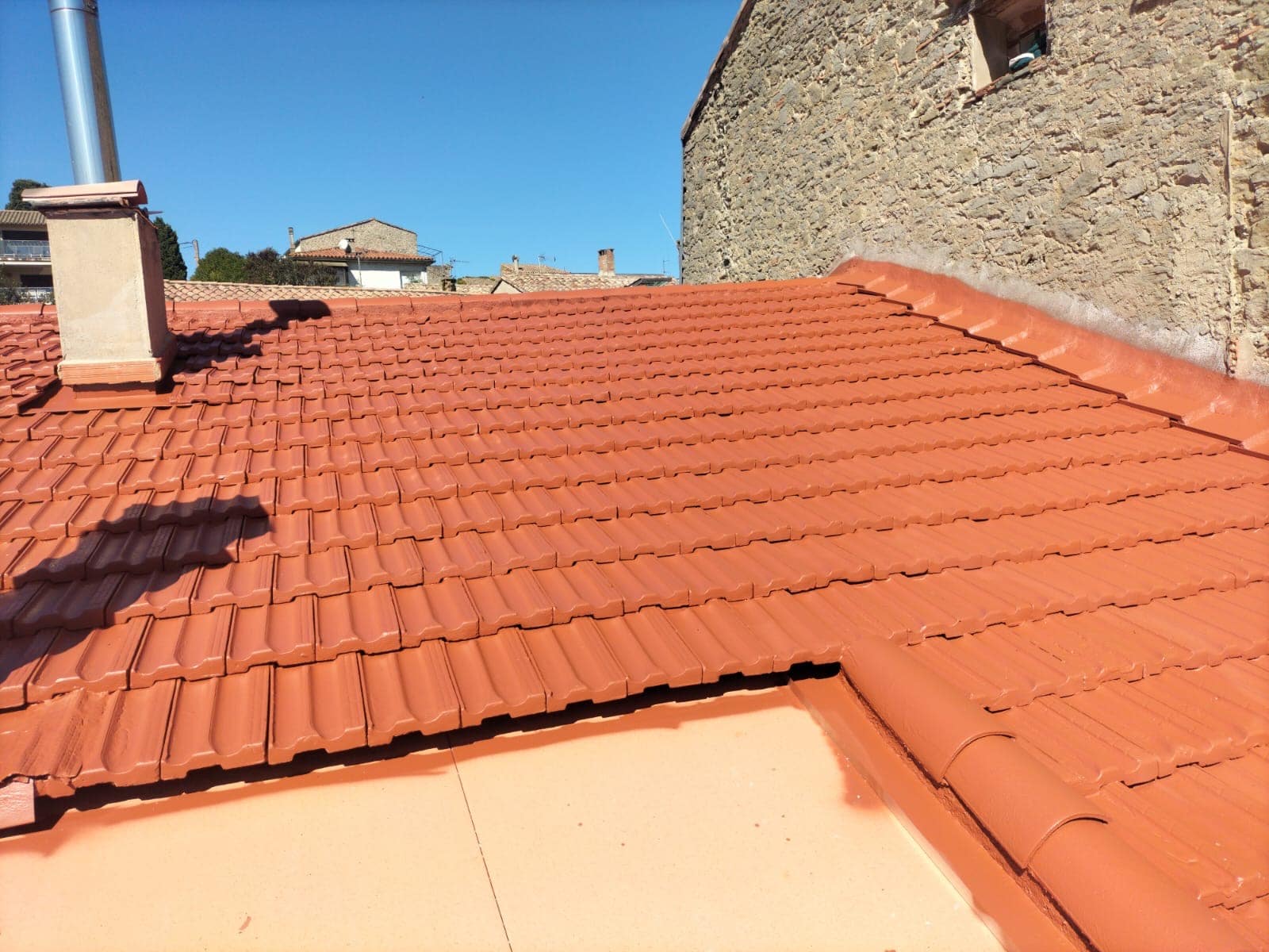 Peinture et Rénovation de Toiture en Tuiles Rouges Toiture en tuiles rouges terre cuite avec cheminée et mur en pierre, rénovation de toiture sous ciel bleu.