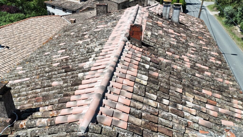 Toit ancien en tuiles canal vieillies avec faîtage et cheminée en brique, vue aérienne dans un village.