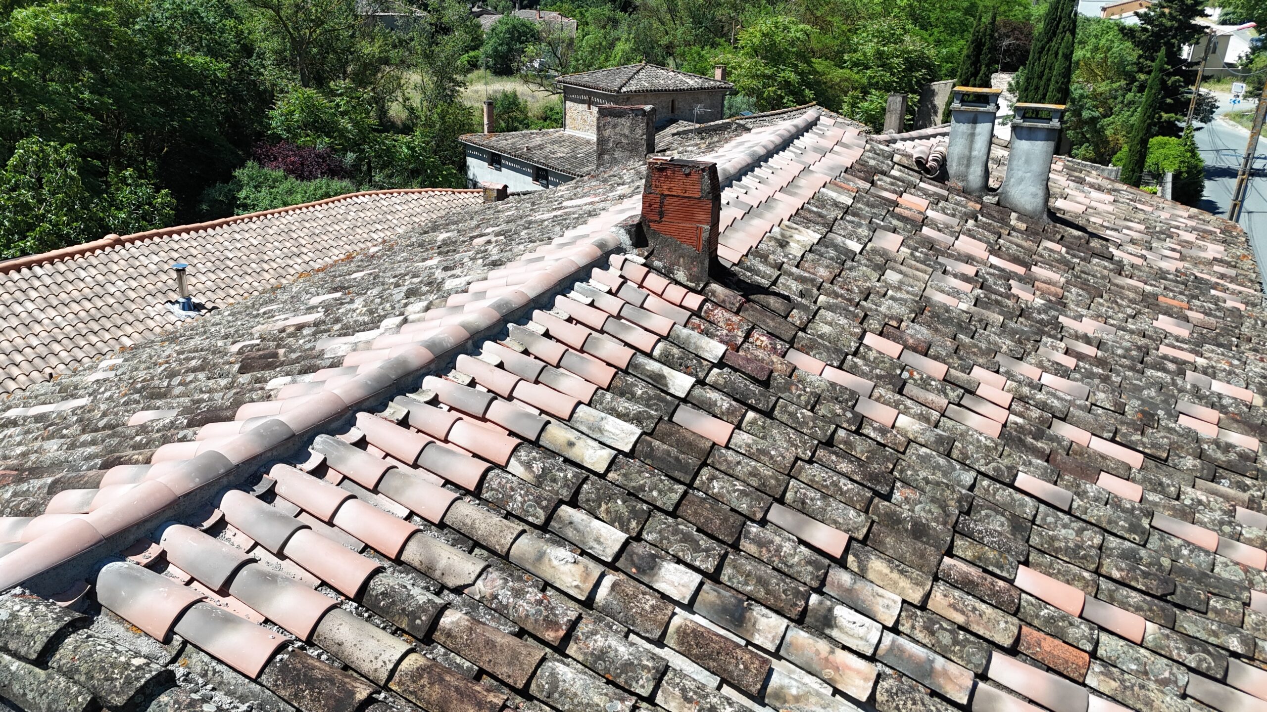 Toit en tuiles romanes vieillies et neuves avec plusieurs cheminées surplombant un village boisé ensoleillé.